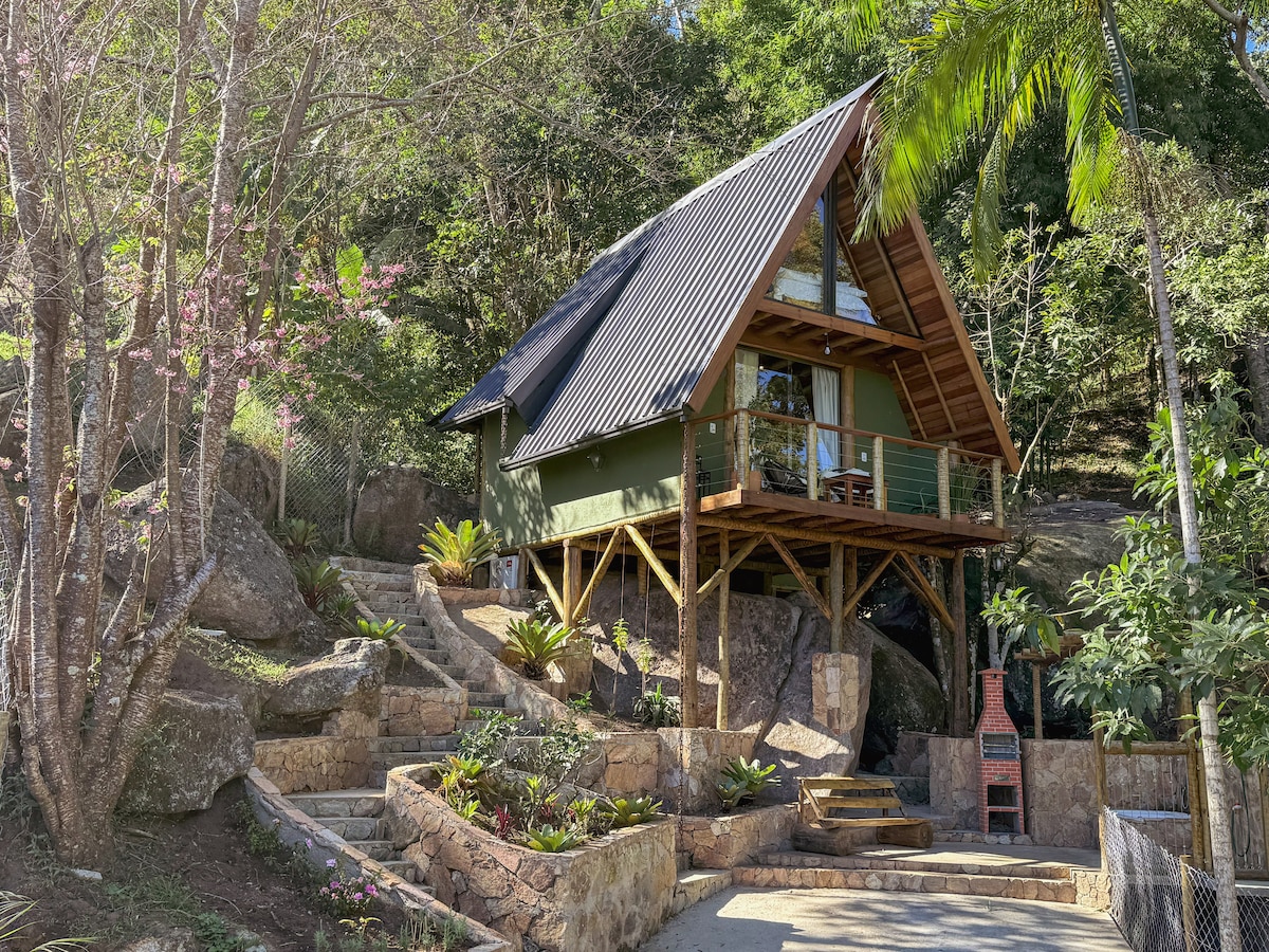A charming chalet is set among lush greenery, featuring a sloped roof and wooden balconies. Steps lead up to the entrance, surrounded by carefully arranged stone pathways and vibrant plants that enhance the natural environment.