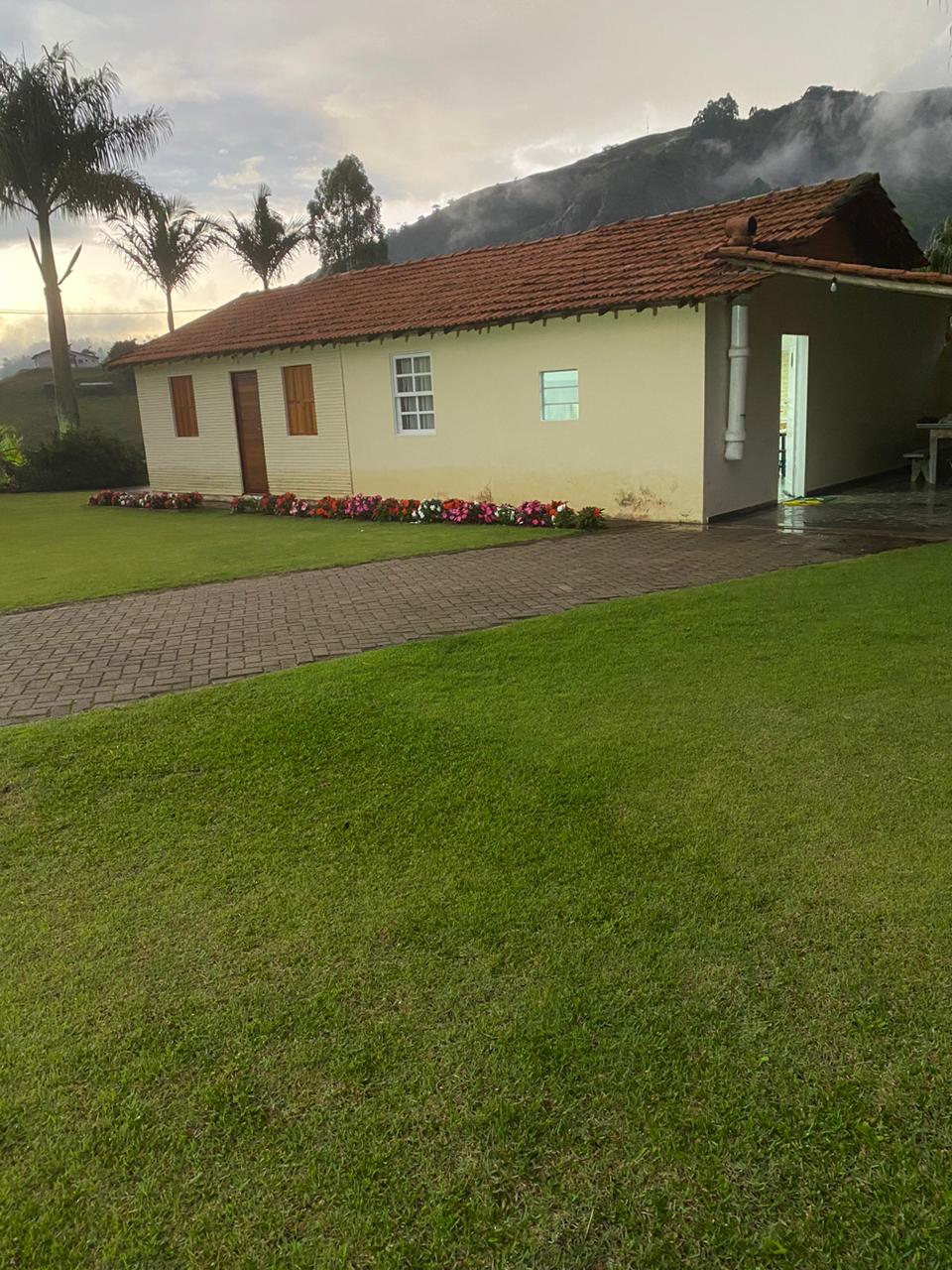 A single-story cottage is shown with a light-colored exterior and a red tile roof. Two wooden doors, along with a window, are present on one side of the building. A lush green lawn surrounds the cottage, with a row of colorful flowers lining the pathway in front.
