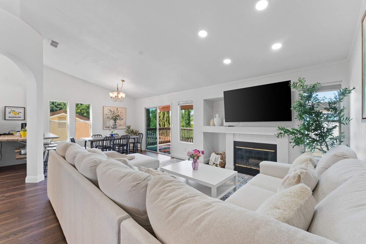 A bright living area features a spacious light-colored sectional sofa facing a modern fireplace. Large windows provide ample natural light and views of the outdoors, while a dining area is visible in the background with a chandelier and a table accommodating multiple guests.