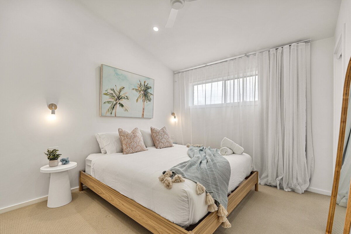 Este dormitorio principal, luminoso y ventilado, ofrece comodidad de estilo resort con una lujosa cama tamaño king, suaves tonos neutros y decoración costera. Las cortinas transparentes, las obras de arte relajantes y la iluminación suave crean un espacio tranquilo para relajarse y recargar energías con estilo.