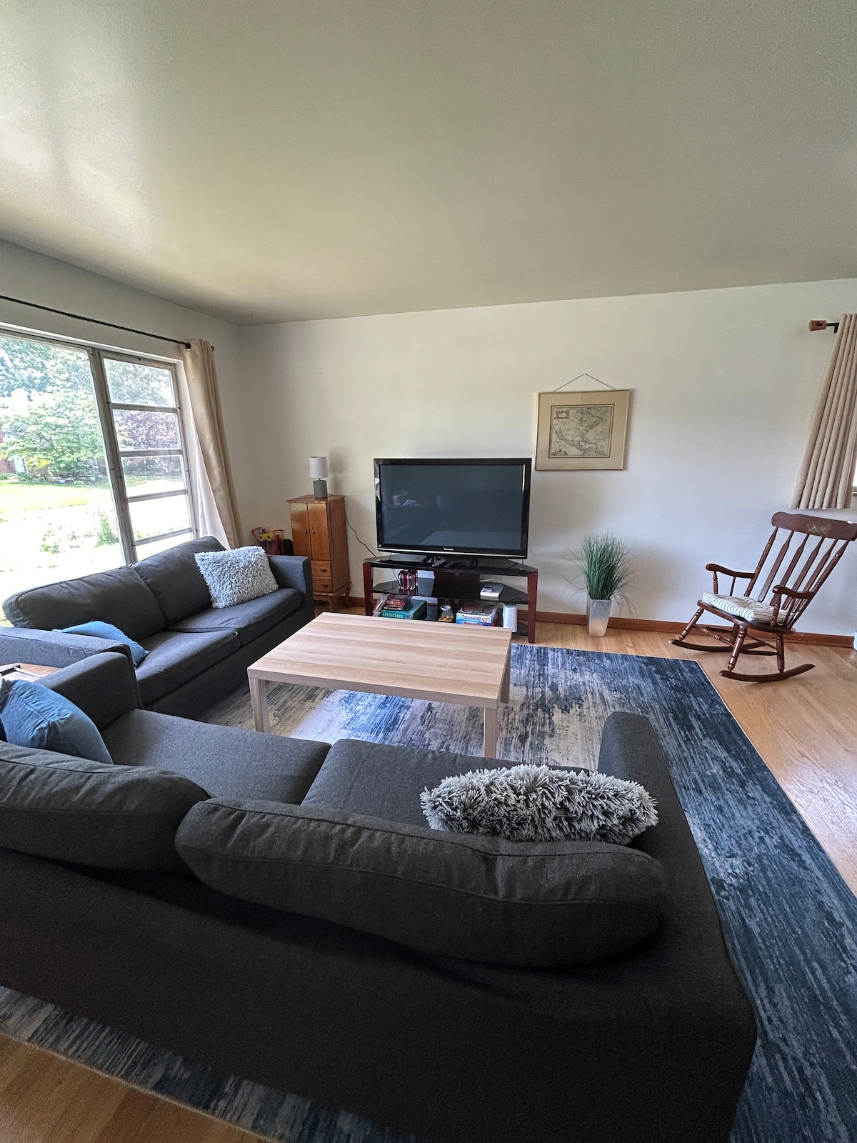 A comfortable living area features two dark sofas and a coffee table, arranged for relaxation. A smart TV stands on a wooden cabinet, while a classic rocking chair is positioned nearby. Large windows provide natural light and views of the backyard.