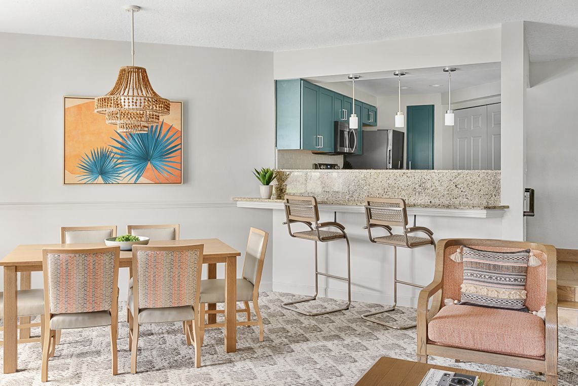 A dining area features a wooden table set for six, complemented by light-colored chairs. A cozy accent chair sits nearby. The walls are adorned with a vibrant artwork, and the open kitchen is visible in the background, showcasing modern appliances and a granite countertop.