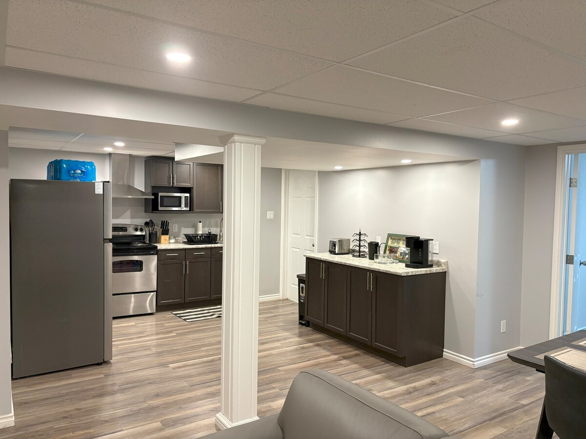 The kitchen area is equipped with modern appliances, including a stove, microwave, and refrigerator. Dark cabinetry contrasts with light countertop surfaces. A small dining area and living space are visible, featuring a neutral color palette and warm lighting from ceiling fixtures.