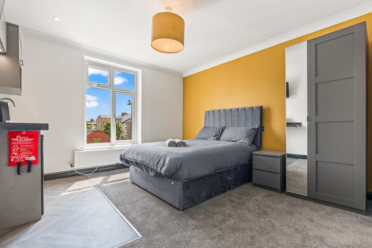 A spacious bedroom is furnished with a large, comfortable bed and a neutral color scheme. A bright yellow accent wall adds a modern touch. Natural light filters through a window, and a stylish light fixture hangs from the ceiling. A bedside table and a mirrored wardrobe complement the space.