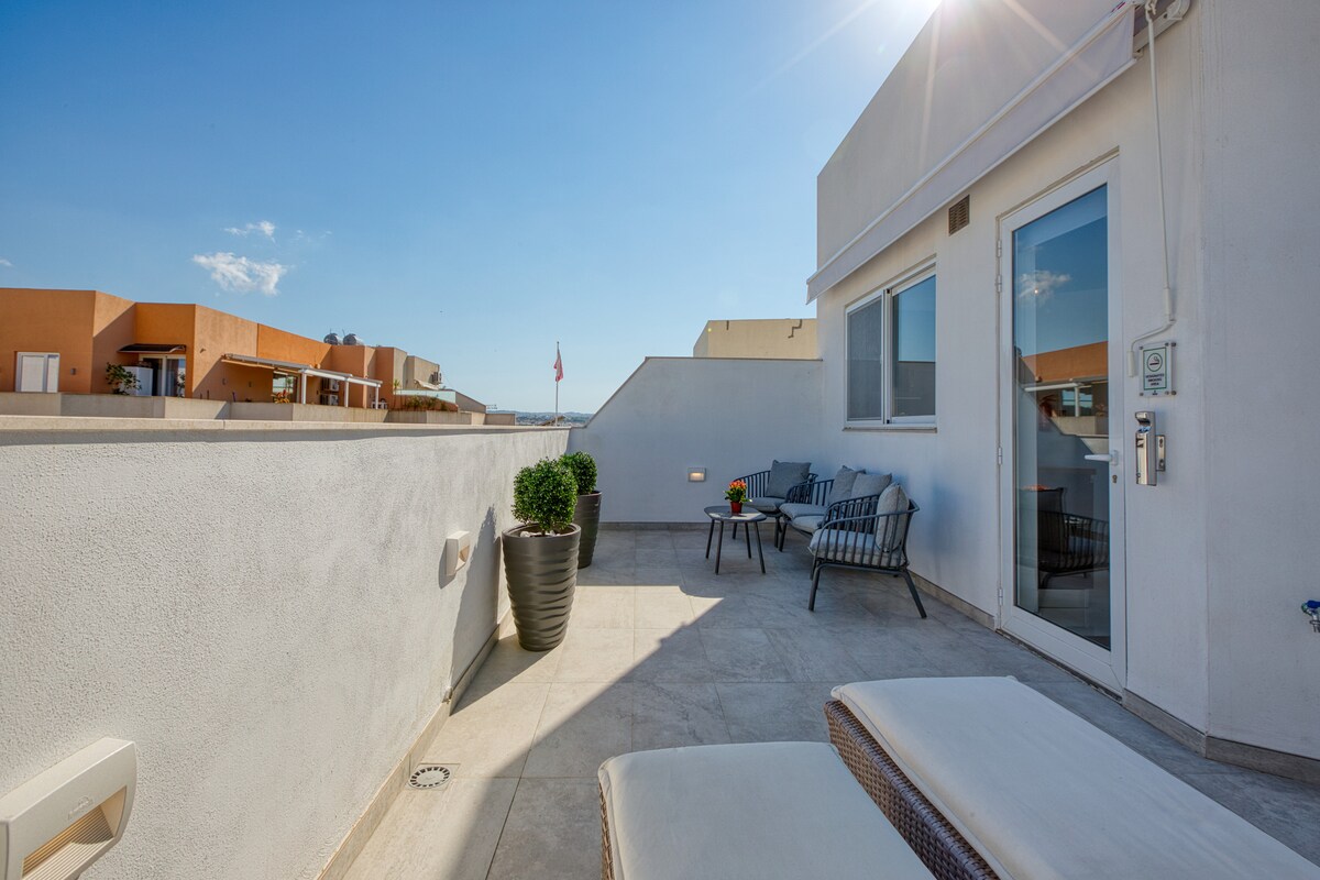 A sunlit private balcony features comfortable seating and a small table, set against a backdrop of clear blue skies. Potted plants add a touch of greenery, while the surrounding wall provides privacy. This outdoor space is ideal for relaxation and enjoying the sunshine.