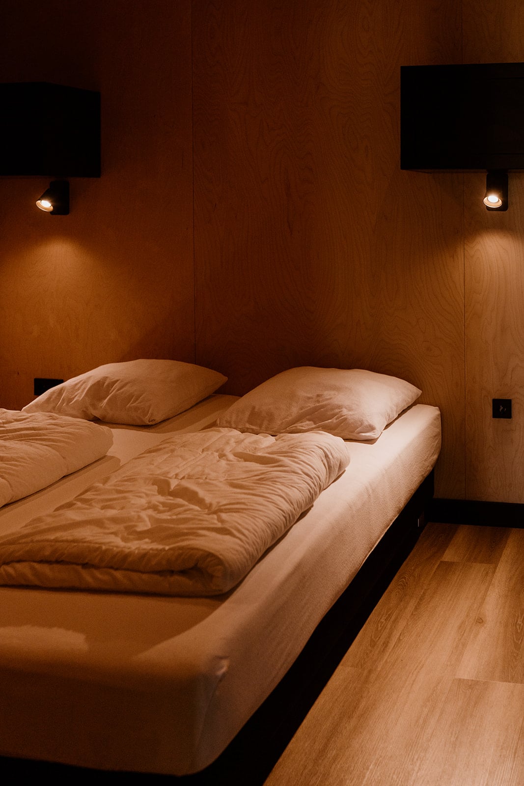 A serene bedroom setting features a spacious bed with two soft pillows and light bedding. Warm wall lights provide a gentle glow against the wooden walls, contributing to a cozy atmosphere. The flooring is adorned with natural wood, enhancing the room's simplicity.