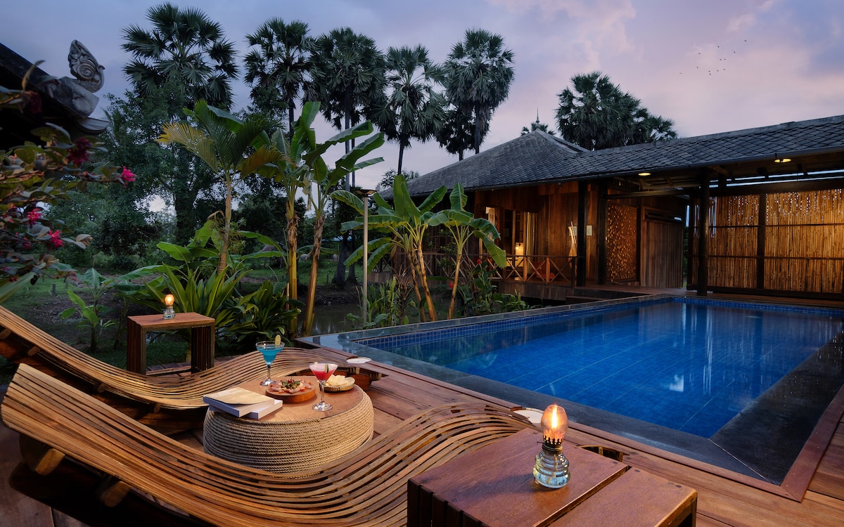 A private pool area is surrounded by tropical plants, featuring comfortable lounge furniture. Soft lighting from lanterns complements the serene blue water of the pool, while trays with snacks and beverages are placed on wooden tables, creating an inviting space for relaxation.