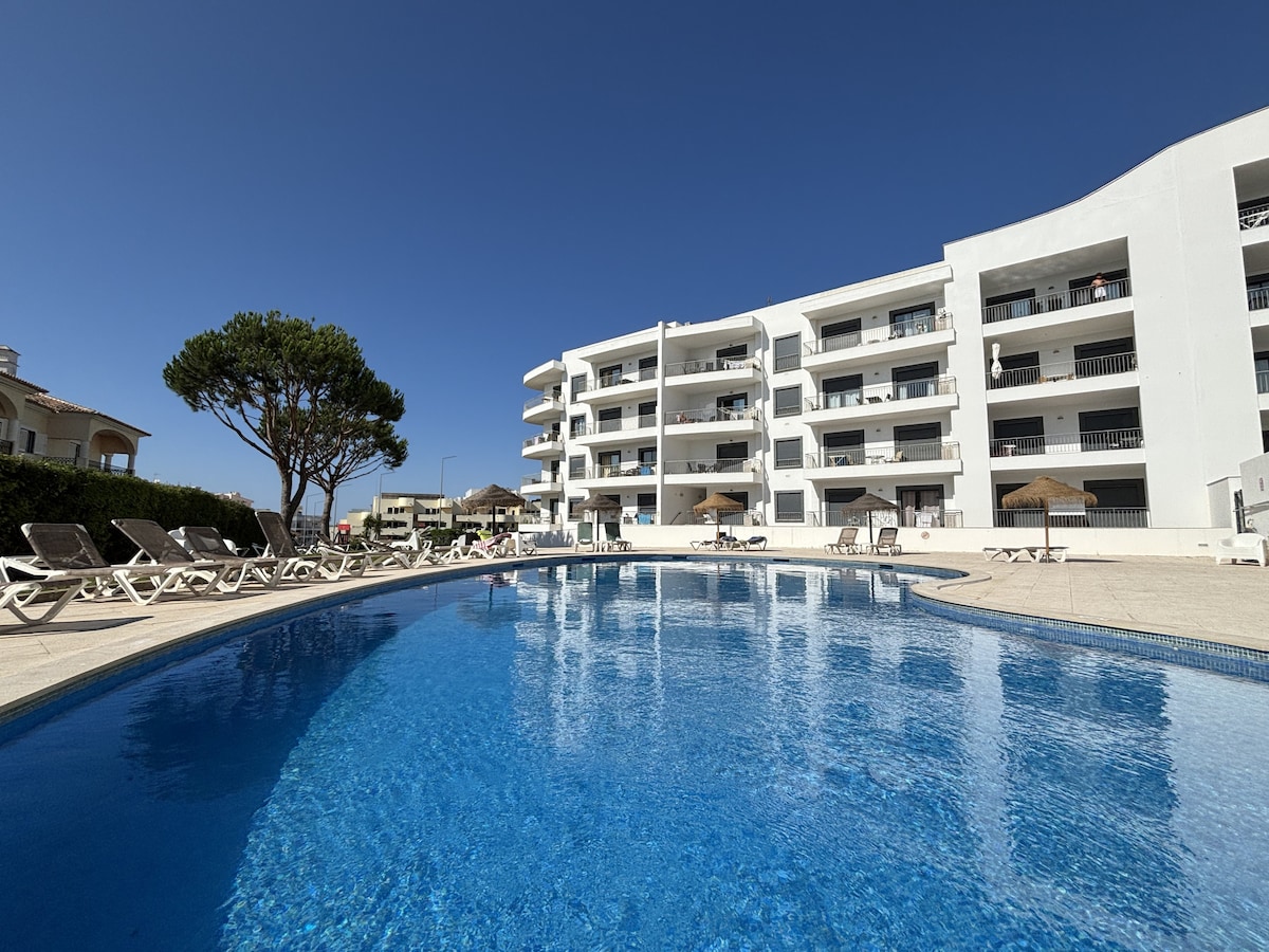 A large swimming pool is surrounded by sun loungers and shaded areas. The pool reflects the sky and is framed by a white residential building. A well-maintained tree stands nearby, offering a touch of greenery to the serene atmosphere.