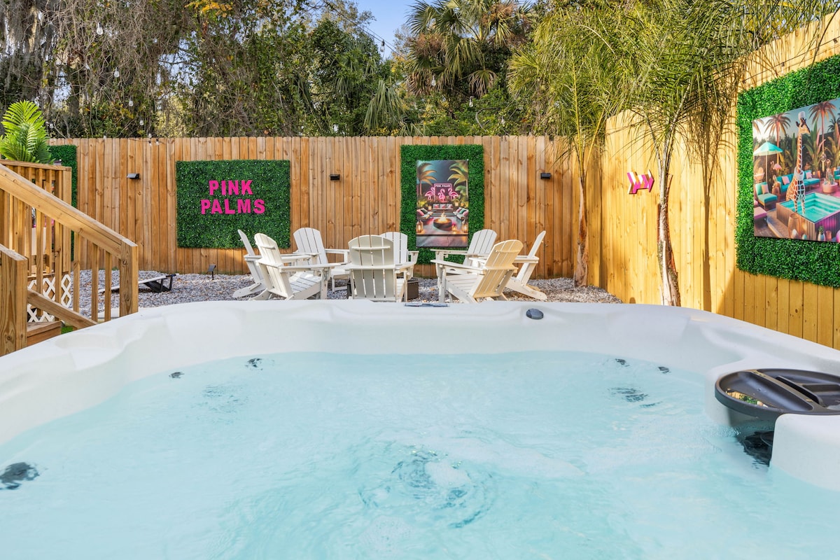 A private hot tub is situated at the forefront, surrounded by a wooden deck. Behind it, a fenced area features a small seating arrangement with four white chairs. Colorful wall art and greenery enhance the space, which is designed for relaxation and enjoyment.