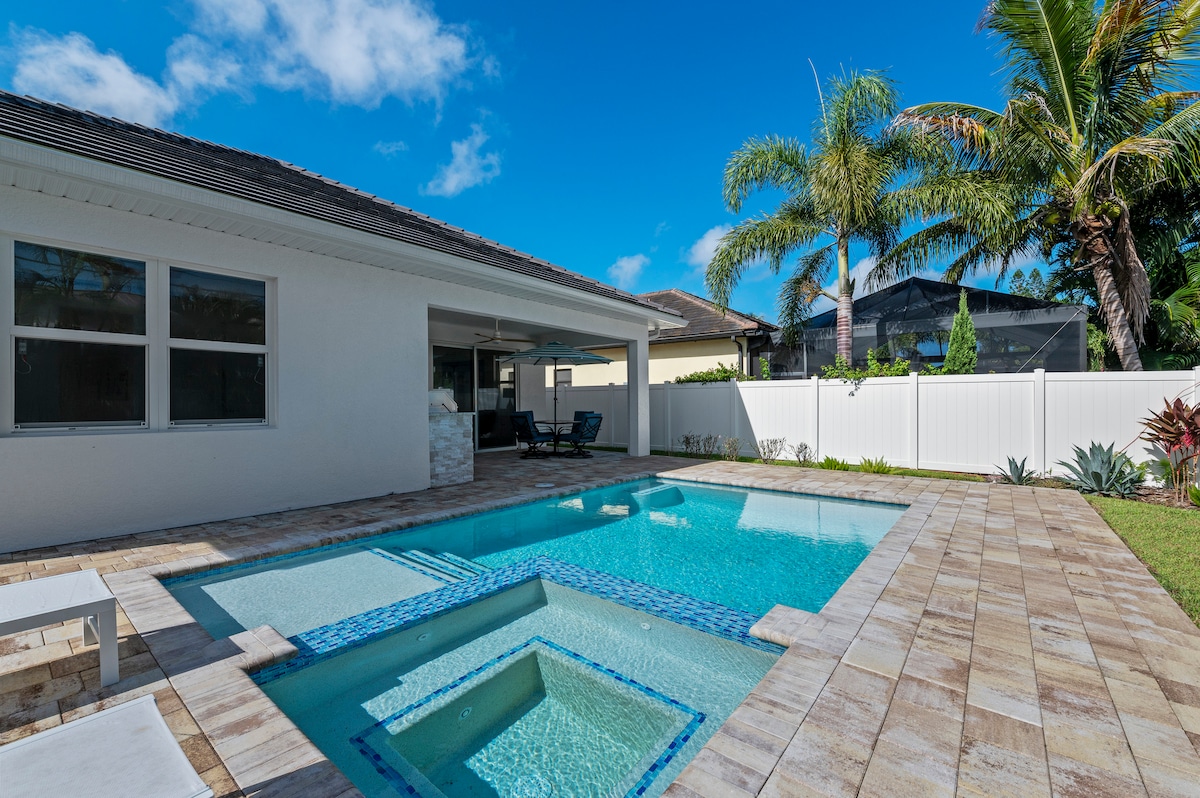 A serene backyard area is highlighted by a tranquil pool with a built-in spa. A spacious patio surrounds the pool, and tropical palm trees can be seen in the background, providing a relaxing outdoor environment.