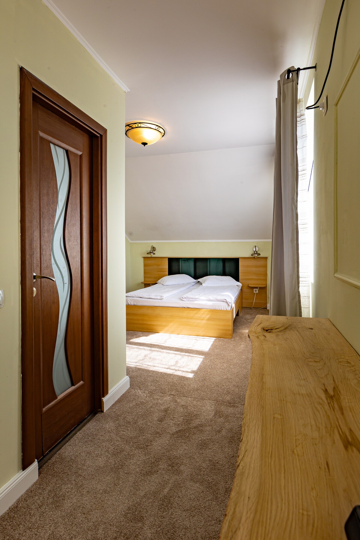 A well-lit hotel room entrance reveals a spacious area featuring a double bed with a light wooden headboard. Soft carpet underfoot complements the pale wall color, while natural light filters through sheer curtains. A decorative wooden console runs along the side wall, enhancing the welcoming atmosphere.