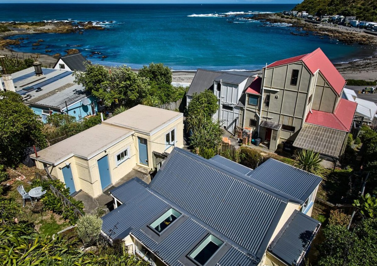 An aerial view showcases the charming seaside cottages nestled along the coast, with the vibrant blue ocean gently lapping at the shore. Lush greenery surrounds the structures, and several rooftops exhibit various shades, contributing to the area's rustic character.