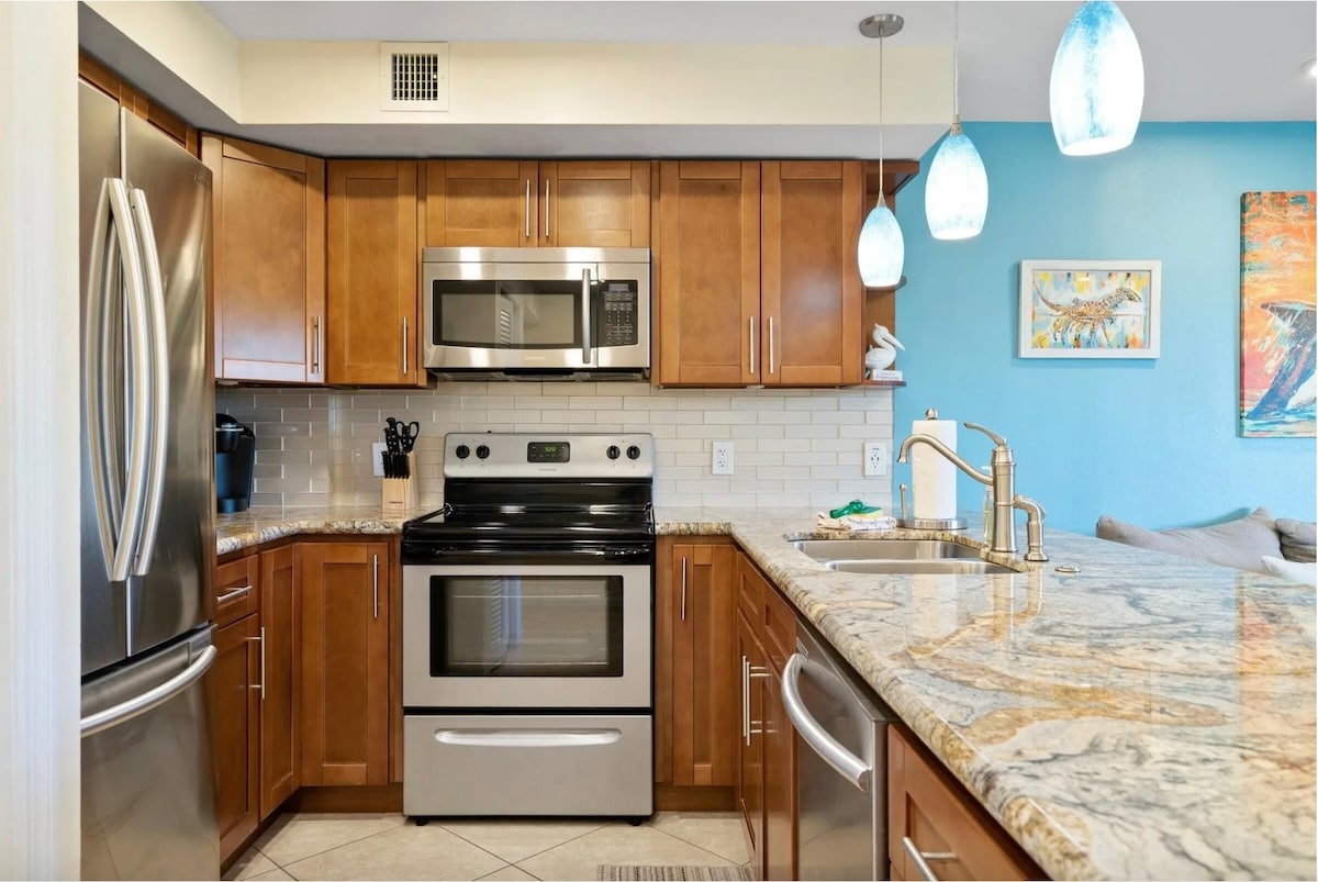 A modern kitchen features warm wooden cabinets and sleek stainless steel appliances. A spacious granite countertop provides ample room for food preparation. Under-cabinet lighting highlights the tile backsplash, while colorful pendant lights offer a pop of color above the counter.