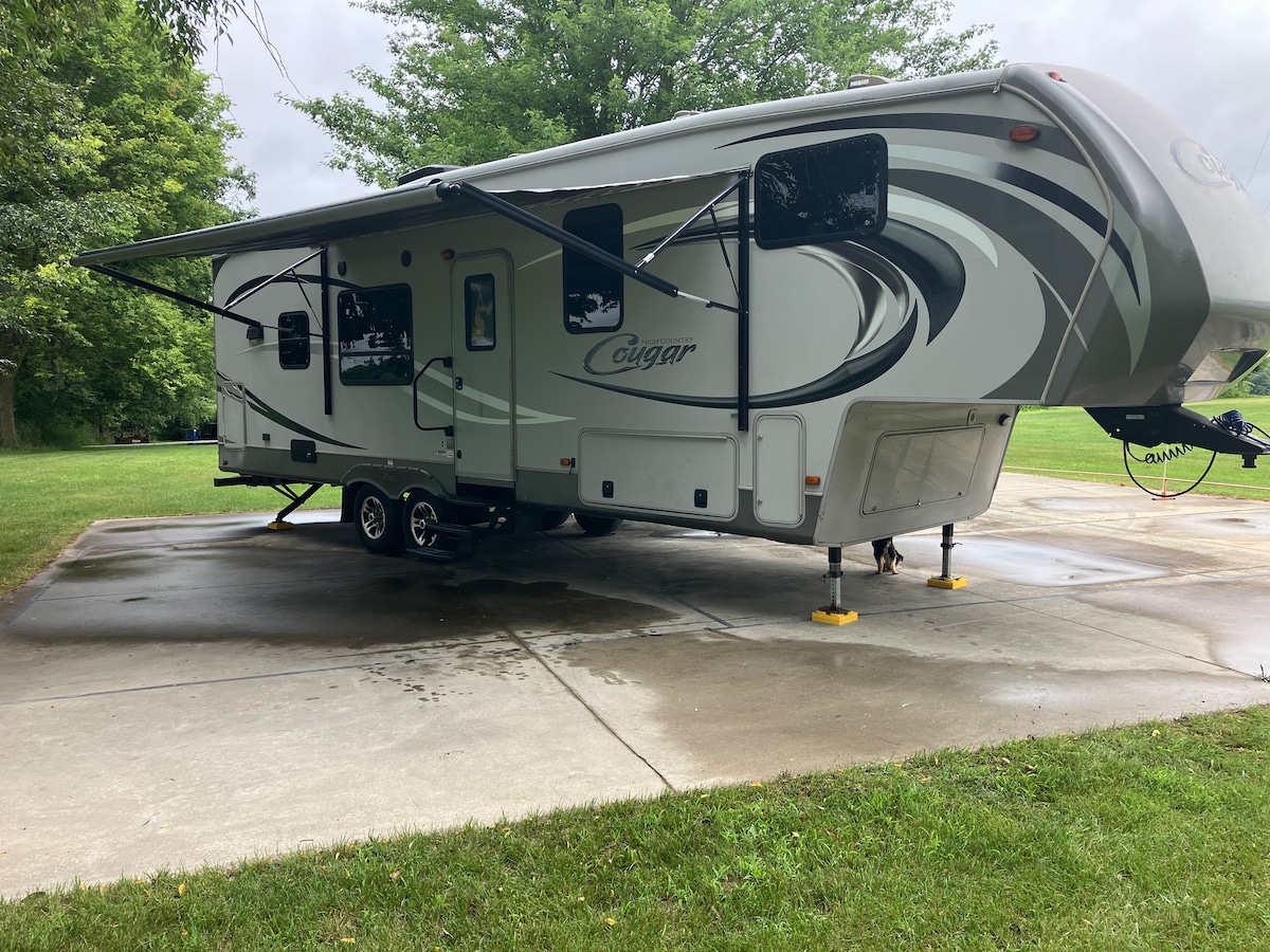 A spacious RV is positioned on a concrete pad, surrounded by lush greenery. A retractable awning extends from the side, providing shade. Nearby, a well-maintained lawn offers an inviting outdoor space for relaxation.