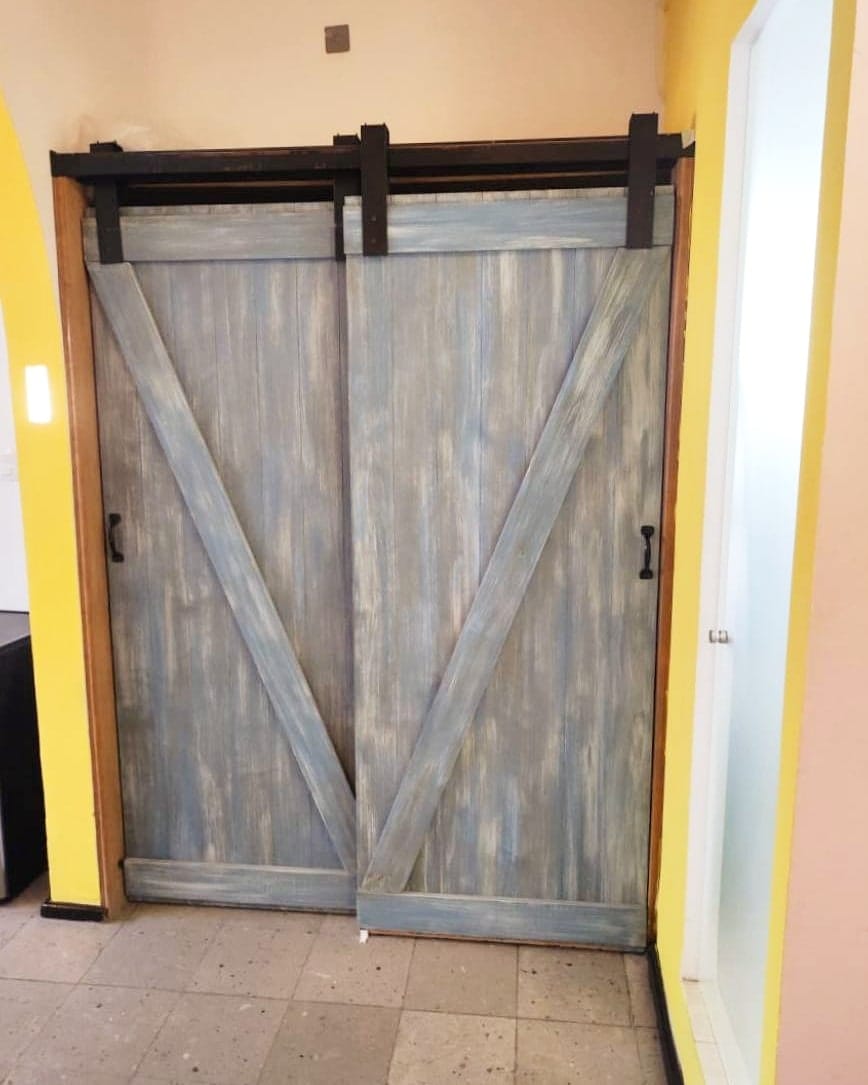A sliding barn door in a rustic finish is mounted on a sleek metal track. The door features a weathered blue hue with diagonal planks, adding character to the room. Adjacent to it, a brightly lit doorway can be seen, framed by a light yellow wall.