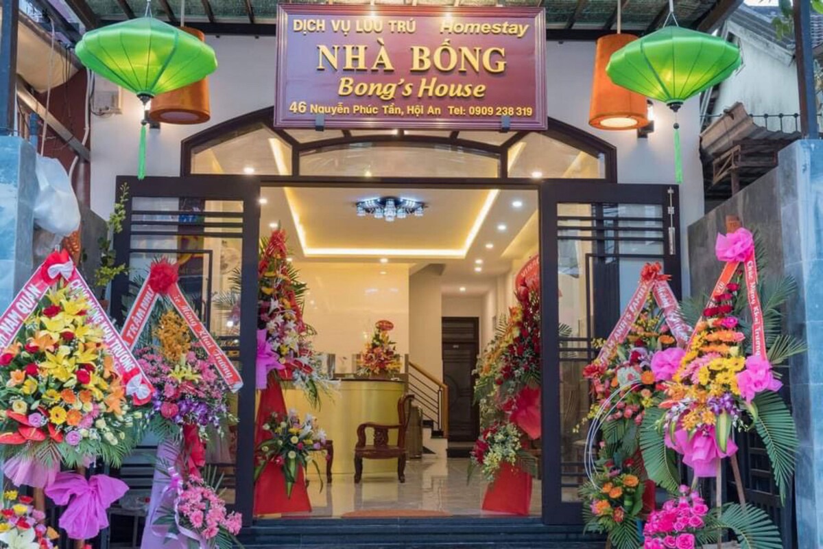 The entrance welcomes guests with vibrant floral arrangements and colorful lanterns. The modern facade features large glass doors leading to a well-lit reception area, with a contemporary desk visible in the background. The overall ambiance is lively, reflecting a friendly hospitality.
