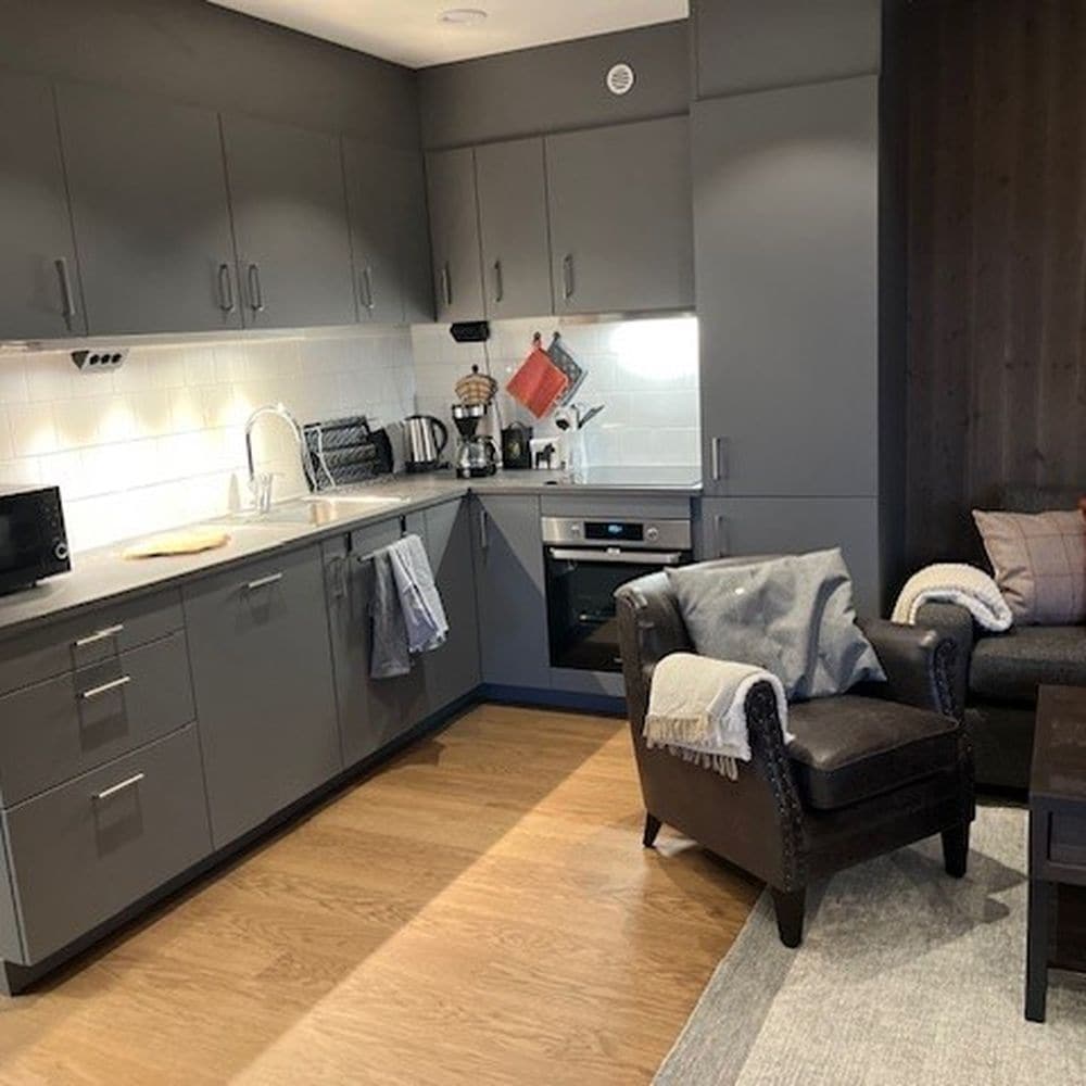 A fully equipped kitchen features modern cabinetry in a subtle grey tone. Essential appliances, including a coffee maker and oven, are easily accessible. A comfortable chair rests nearby, with a cozy throw draped over its arm, complementing the warm wood flooring.