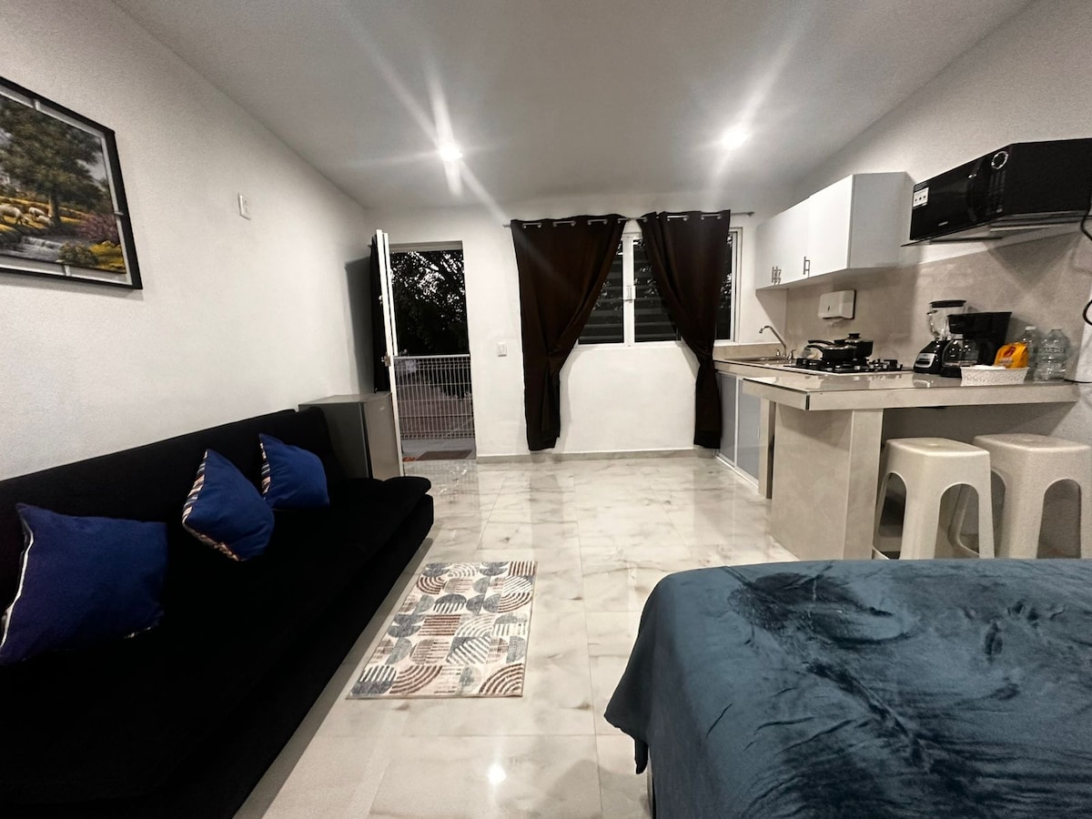 A well-arranged living space is shown featuring a black sofa adorned with decorative pillows. A compact kitchen is visible, equipped with essential appliances. Light-colored tiles enhance the overall brightness, while dark curtains frame the window, and a small rug adds texture to the floor.