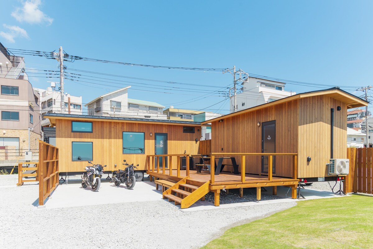 A wooden structure features a spacious deck with seating options and two motorcycles parked in front. The exterior showcases large windows, allowing ample natural light. The surrounding area includes a well-maintained gravel space and a lawn, contributing to a serene outdoor atmosphere.