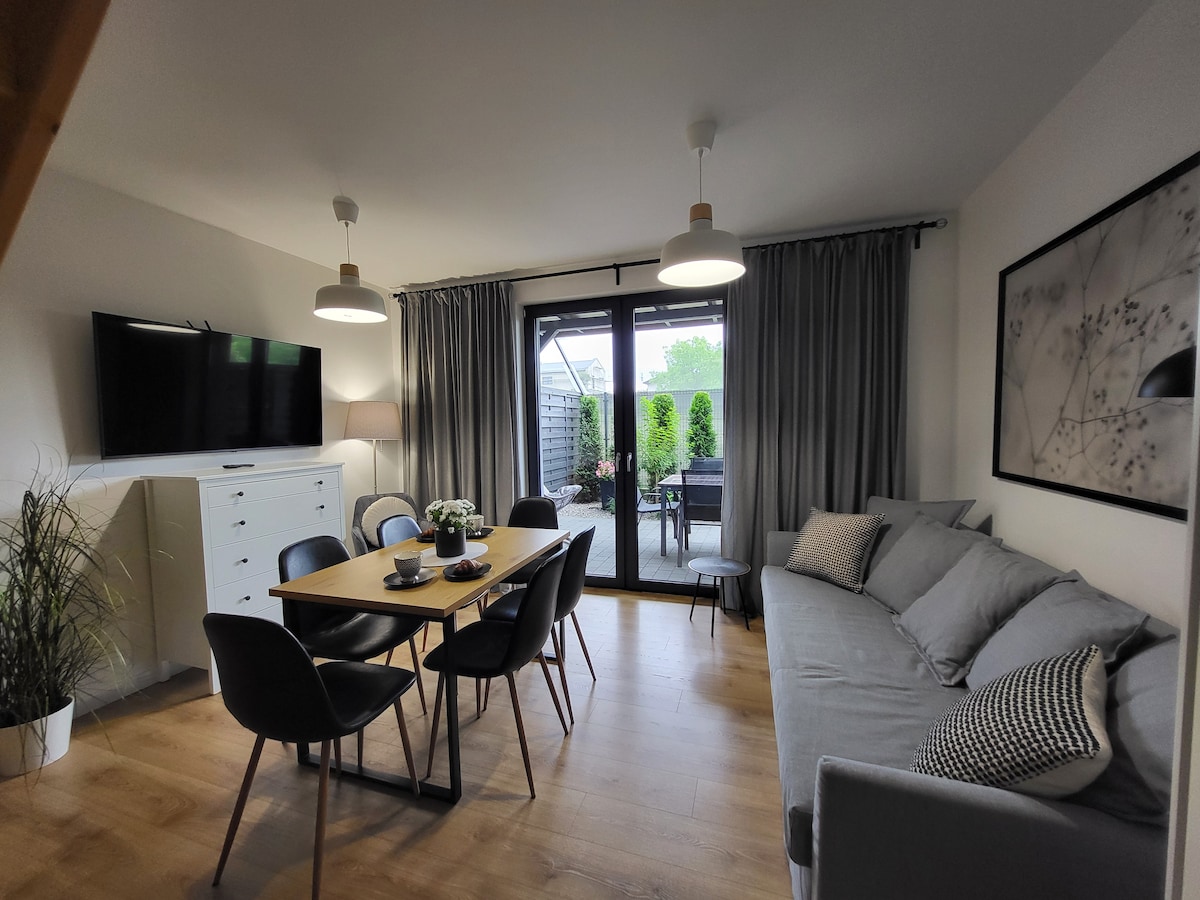 A modern living area is presented with a spacious sofa and a wooden dining table surrounded by black chairs. Large glass doors open to an outdoor space, while neutral-toned walls and wooden flooring create a harmonious atmosphere. Soft lighting is provided by ceiling fixtures.