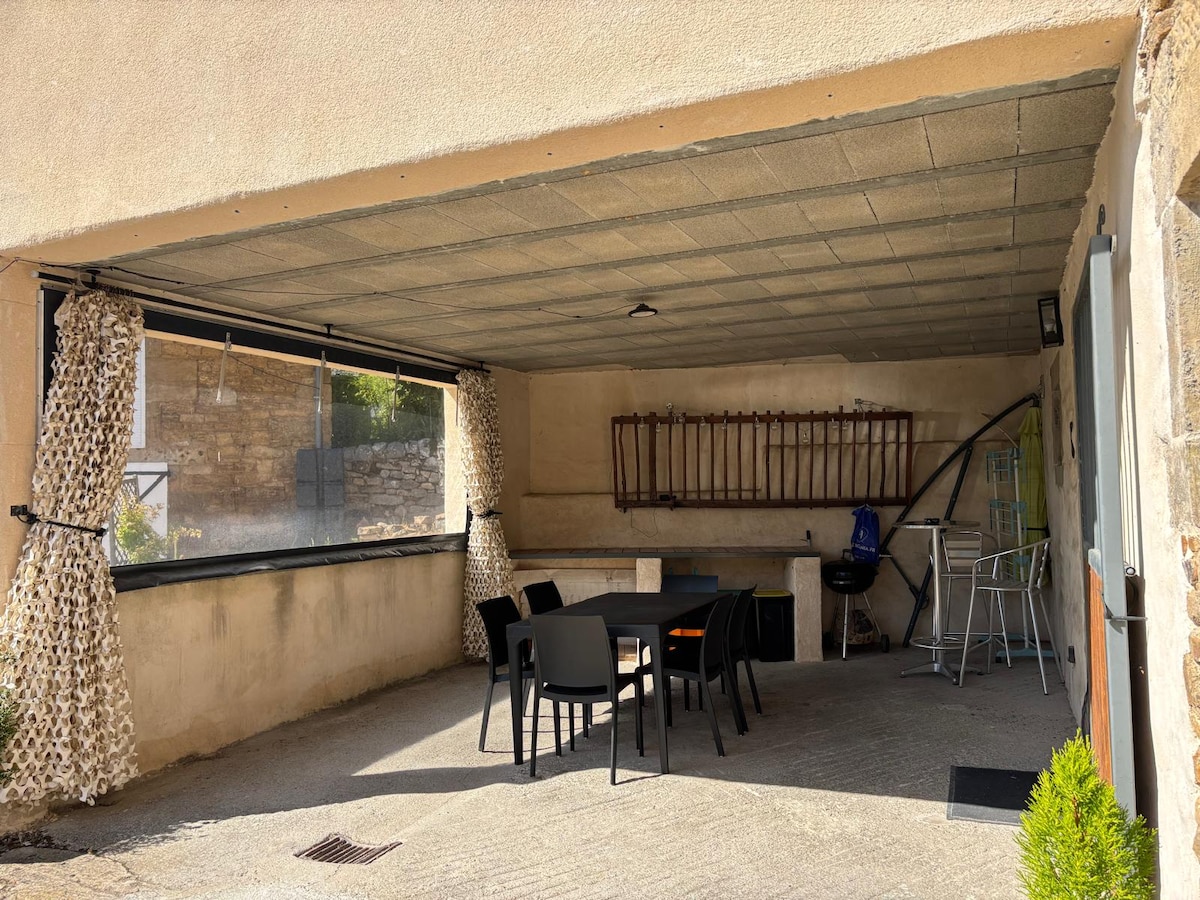 A covered outdoor space features a table with six black chairs, ideal for dining or social gatherings. A charcoal barbecue is positioned nearby, with a drying rack and additional storage area visible on the wall. Natural light enters through the open sides.