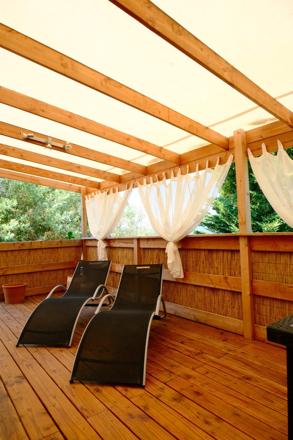 A spacious wooden terrace features two shaded lounge chairs positioned for relaxation. Light curtains hang elegantly, providing a sense of privacy while natural greenery is visible in the background, enhancing the outdoor setting.