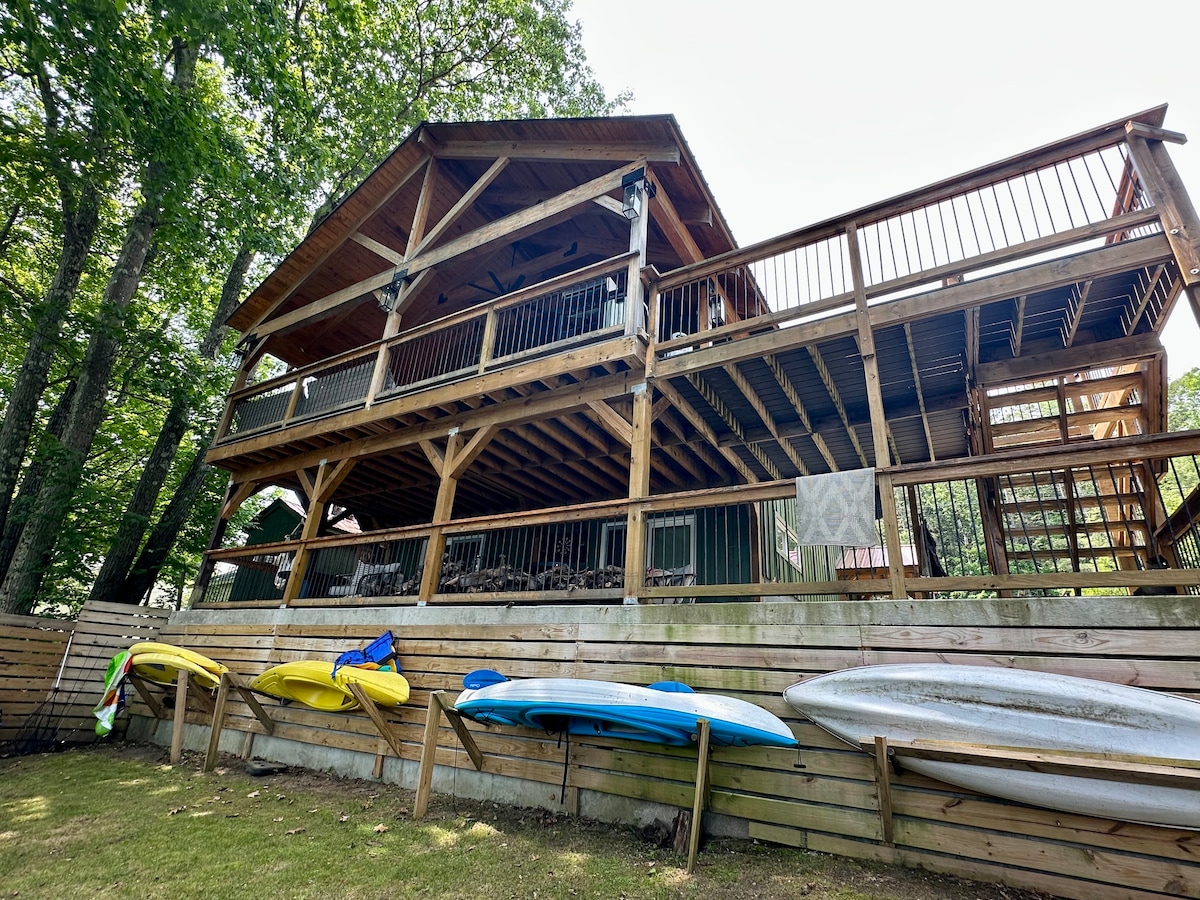The exterior of a two-story wooden structure is surrounded by trees. A spacious covered deck is visible on the upper level, featuring railings and overhead lights. Kayaks are mounted on a wooden support against the lower level, providing convenient lake access.