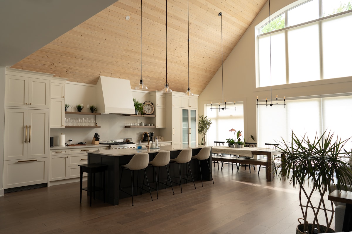 An airy kitchen and dining area feature high ceilings with wooden beams and large windows allowing natural light to fill the space. A central island with seating offers a functional gathering spot, while potted plants add a touch of greenery to the neutral decor.