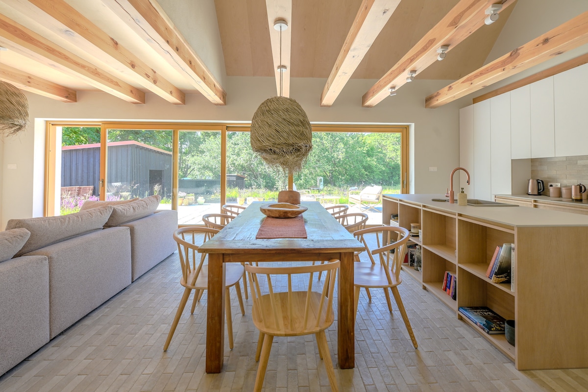 A spacious dining area features a long, wooden table surrounded by light-colored chairs, offering ample seating. Natural light streams through large glass doors, opening to a lush outdoor view. The room's wooden beams contribute to a warm ambiance, complemented by simple decorative elements.