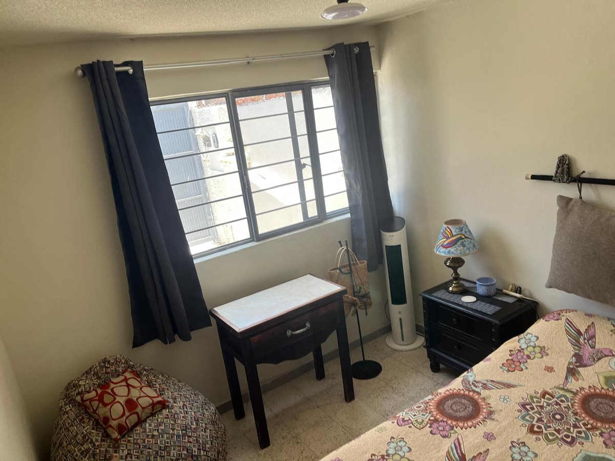 A bright bedroom is illuminated by natural light streaming through a large window, adorned with dark curtains. A patterned bedspread covers the spacious bed, while a small table and decorative lamp sit beside it. A cozy chair adds to the inviting space.