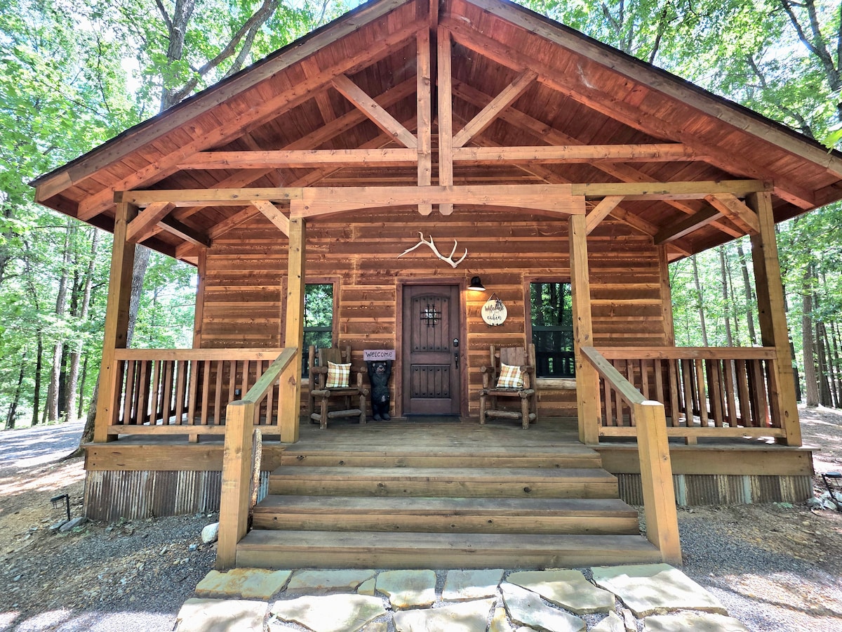 The cabin's inviting entrance features a wooden porch with two rustic chairs and decorative cushions. The structure showcases a combination of log design and a stone pathway, framed by green foliage. A wooden door with a decorative sign and antlers accentuates the welcoming atmosphere.