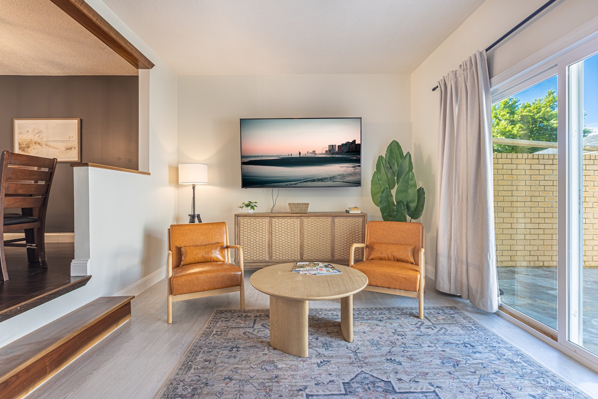 A living area features two modern orange chairs positioned beside a round wooden coffee table. A large 75" 4K Smart TV is mounted on the wall above a woven cabinet, facing a large window that allows natural light to brighten the space. A potted plant adds a touch of greenery.