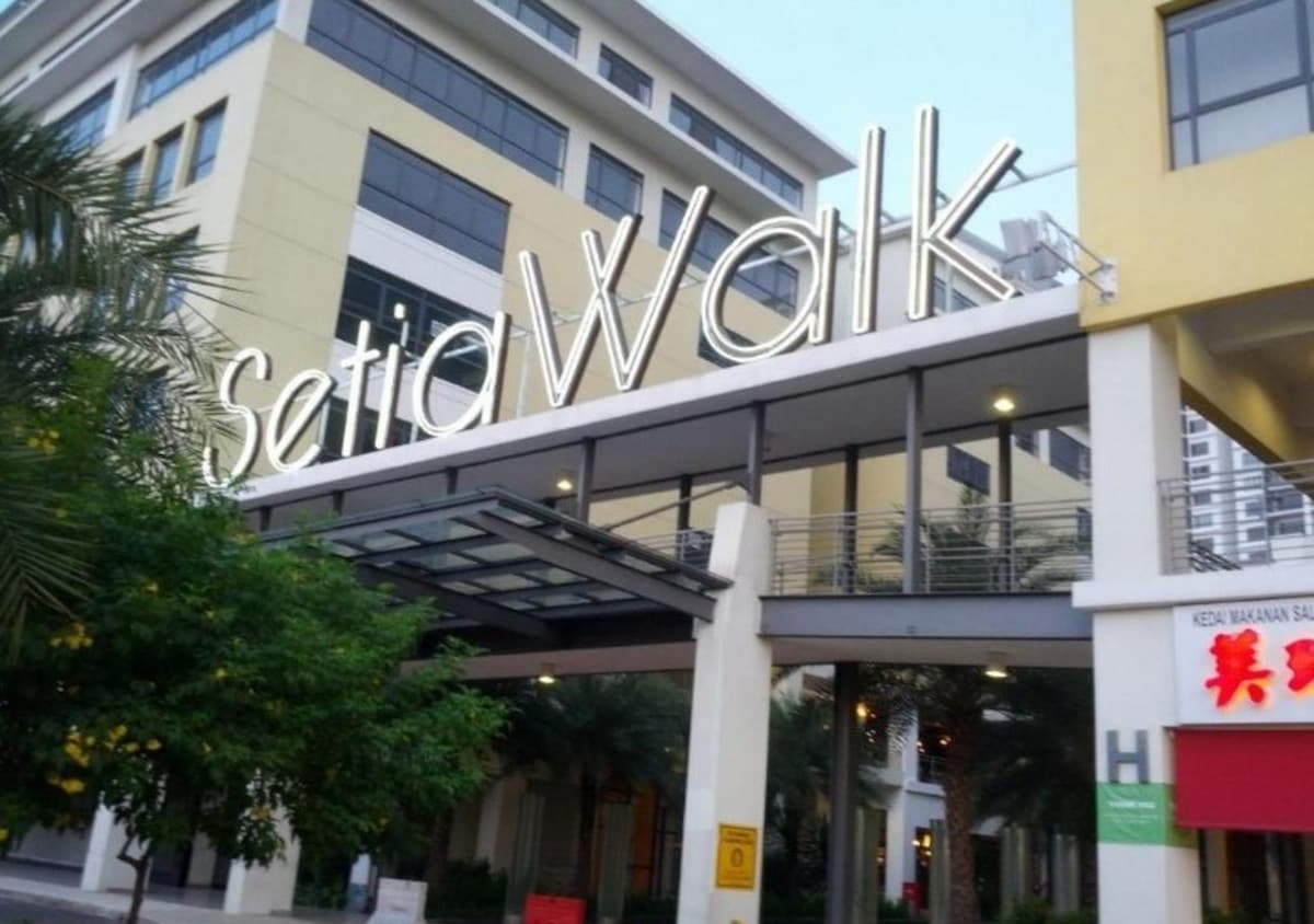 The image features the modern entrance of SetiaWalk, characterized by large signage that reads 'setiaWalk.' The architecture includes sleek buildings and a covered walkway. Lush greenery is visible in the surroundings, contributing to the vibrant atmosphere of the area.