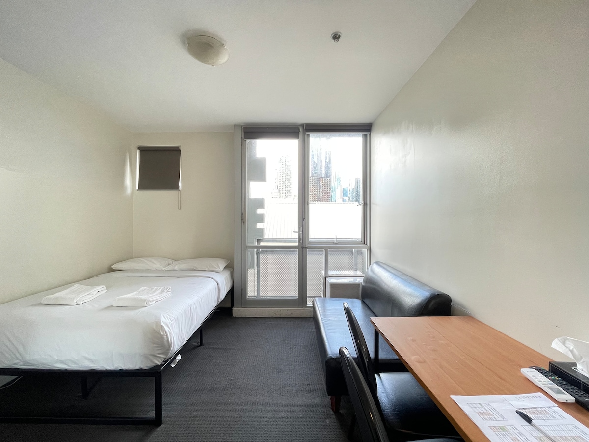 An open layout studio is shown, featuring a spacious bed with fresh linens. A large window offers natural light and city views. An adjacent black leather sofa sits beside a wooden table, creating a comfortable seating area. Storage and a desk are available in the background.