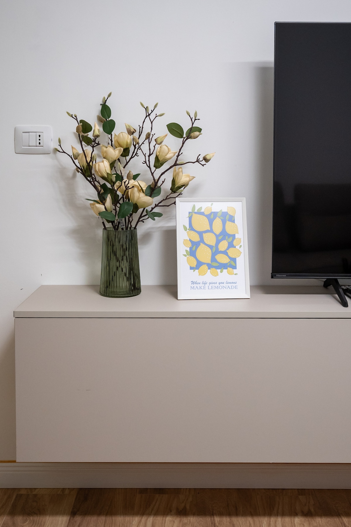 A minimalist entertainment unit is shown, featuring a flat-screen television and a decorative vase filled with artificial flowers. Next to the vase, a framed piece of art adds a pop of color, showcasing a modern design against the neutral backdrop.