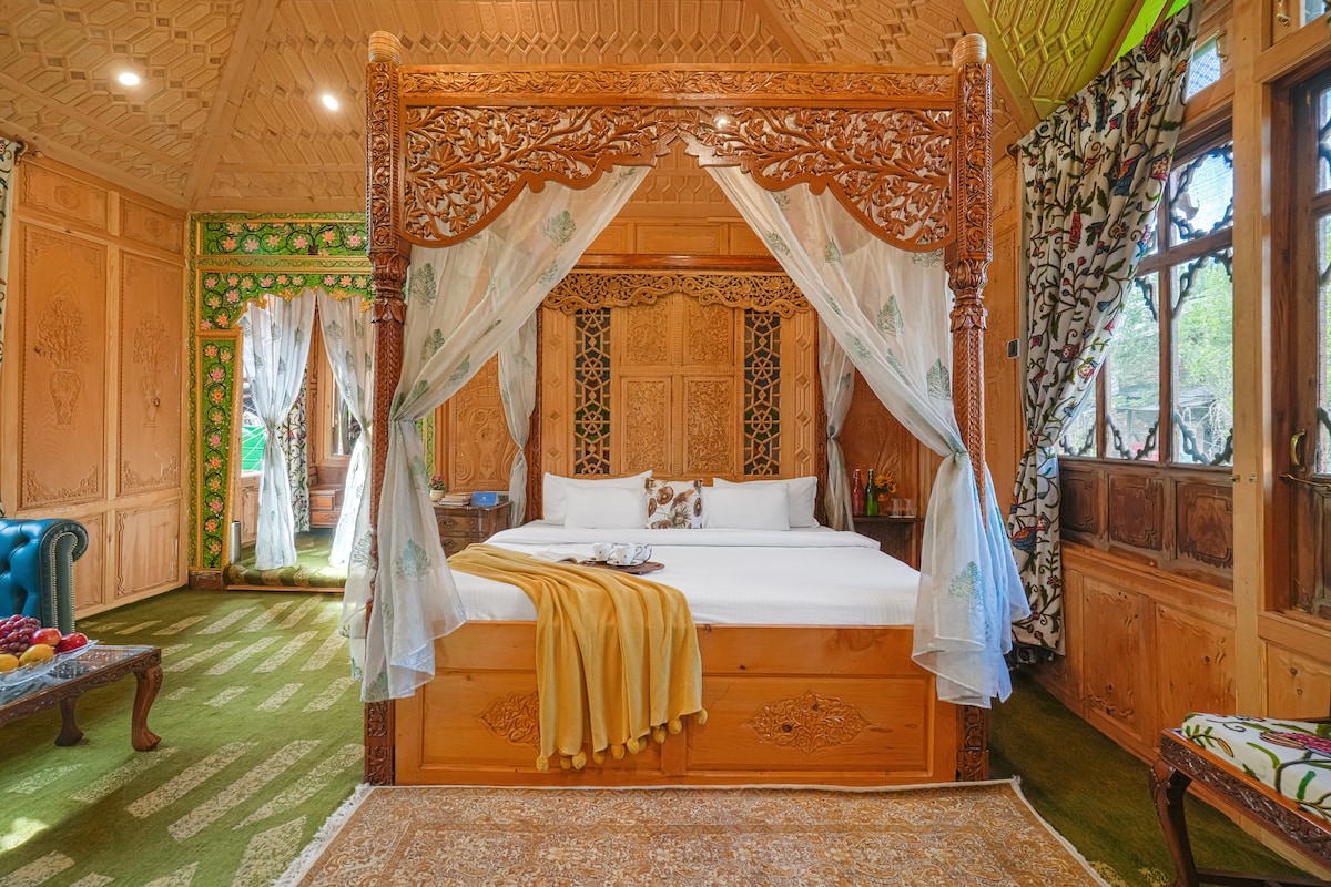 The Shareen Suite is highlighted by an intricately carved wooden four-poster bed, adorned with sheer curtains. Surrounding walls exhibit traditional Kashmiri woodwork, while bright floral patterns on drapes and a vibrant green carpet add charm. Natural light streams through multiple windows, enhancing the inviting space.