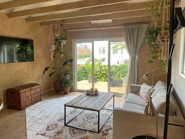 A comfortable living area features a light-colored sofa complemented by a wooden coffee table. Plants hang from the ceiling and decorate the space, while a flat-screen TV is mounted on the wall. Large glass doors open onto a deck, allowing natural light to fill the room.