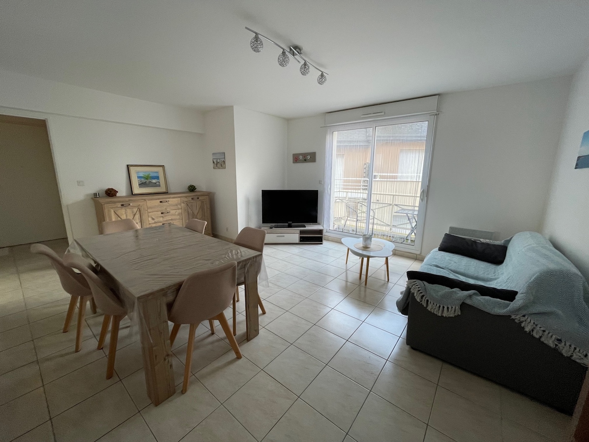 A light-filled living area features a wooden dining table with six chairs and a gray couch. A flat-screen television is positioned on a low stand, while large sliding glass doors lead to an outdoor balcony, allowing natural light to brighten the space.