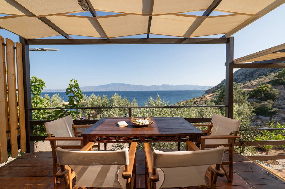 An outdoor dining area features a wooden table accompanied by four light-colored chairs. A large awning provides shade, while a serene view of the sea and hills can be seen in the background. Vegetation adds a natural touch to the setting.