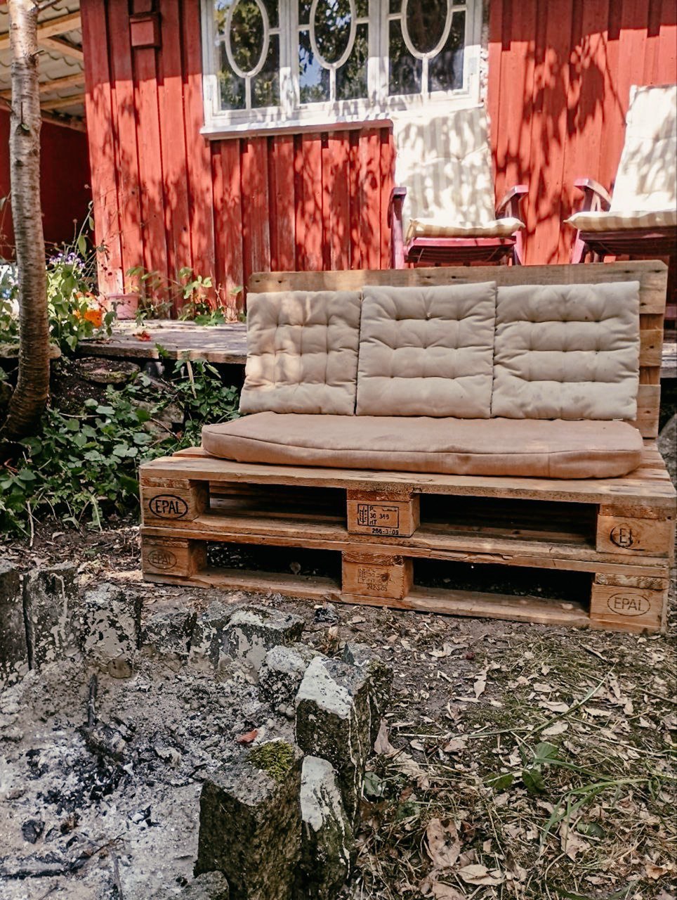 An outdoor seating area features a cushioned couch made from wooden pallets, positioned next to a firepit. The surrounding garden is lush with greenery and the rustic cabin with red walls stands in the background. Sunlight filters through the trees, creating a serene atmosphere.