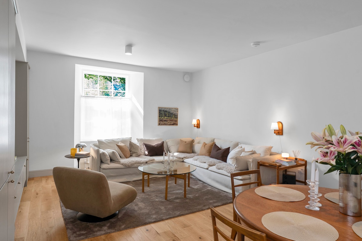 A spacious living area is showcased, featuring a comfortable sectional sofa adorned with neutral cushions. A round dining table with wooden chairs is positioned nearby. Natural light filters through a large window, illuminating a decorative coffee table and a subtle floral arrangement.