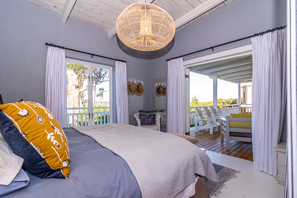 A spacious bedroom is illuminated by natural light coming through large windows. A comfortable bed is layered with neutral-toned bedding, while a decorative throw pillow adds a pop of color. A unique woven light fixture hangs from the ceiling, and seating is visible on the balcony.