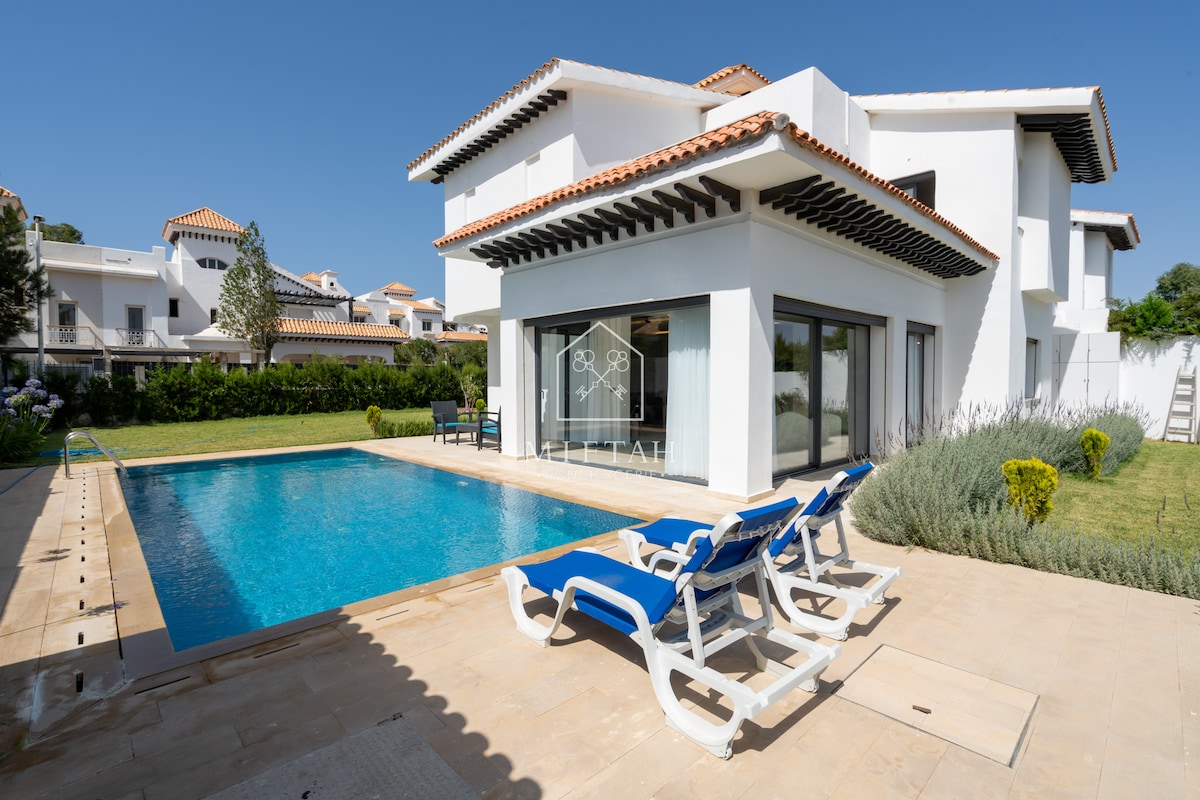 A modern villa exterior is showcased, featuring a spacious swimming pool surrounded by sun loungers. Lush greenery lines the pool area, while large glass doors provide access to the interior. The villa is designed with white and terracotta elements against a clear blue sky.