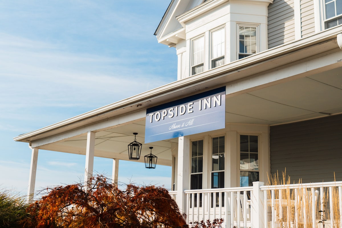 The exterior of a charming inn is highlighted, featuring a large sign that reads 'Topside Inn.' A covered porch with two hanging lanterns provides a welcoming entrance. Lush foliage and tall grass frame the entrance, while the building showcases multiple large windows that create an inviting facade.