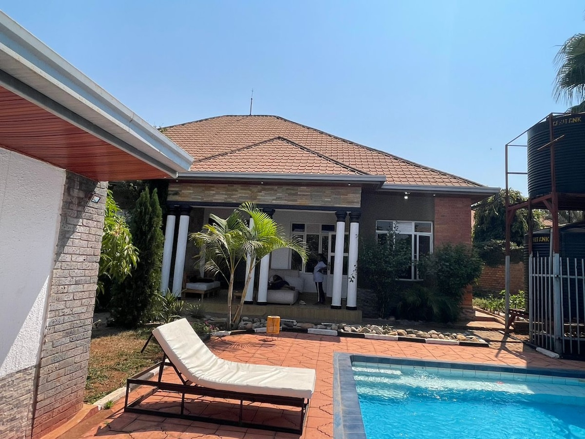 A spacious homestay is depicted, featuring a large swimming pool alongside a sun lounger. The structure showcases an open porch area with multiple seating arrangements and is surrounded by lush greenery. Clear skies are visible above, enhancing the outdoor atmosphere.