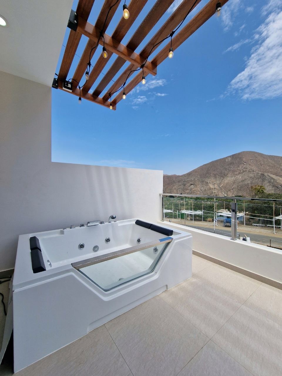A private outdoor space features a modern hot tub, positioned against a backdrop of dramatic mountains. Natural light fills the area, accented by a wooden pergola overhead with decorative string lights, creating a serene setting for relaxation.