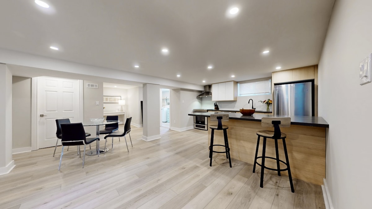 A modern kitchen and dining area is presented, featuring sleek countertops and stainless steel appliances. A dining table with four chairs sits adjacent to the kitchen, while two bar stools are positioned at the counter. The space benefits from ample lighting and a spacious, open layout.
