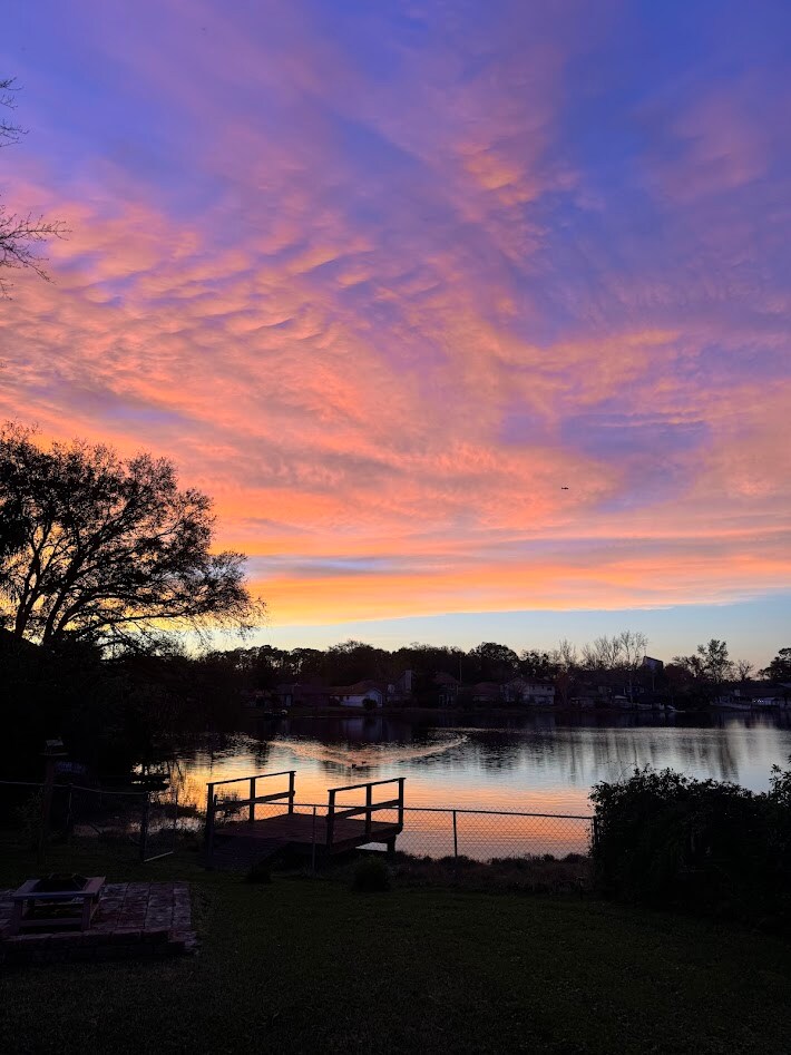 A serene lake view is captured during twilight, showcasing a vibrant sky filled with hues of orange, pink, and purple. A wooden dock extends toward the water, framed by a grassy area and mature trees, enhancing the tranquil natural setting.