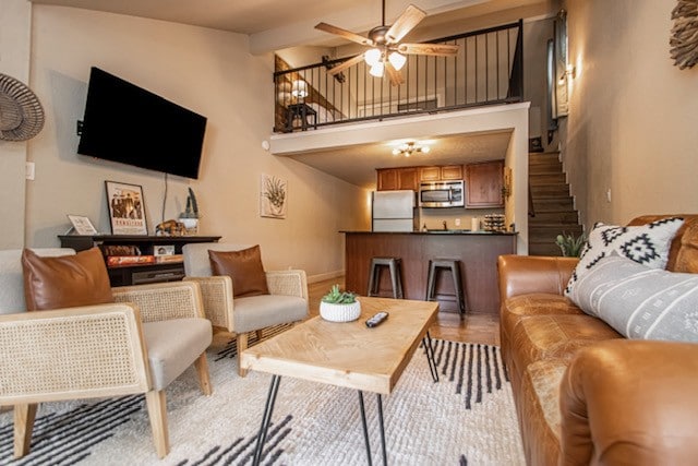 A living area features a warm color palette with a brown leather sofa and two light-colored armchairs. A wooden coffee table sits in the center, surrounded by a textured area rug. A television is mounted on the wall, and a staircase leads to an upper level.