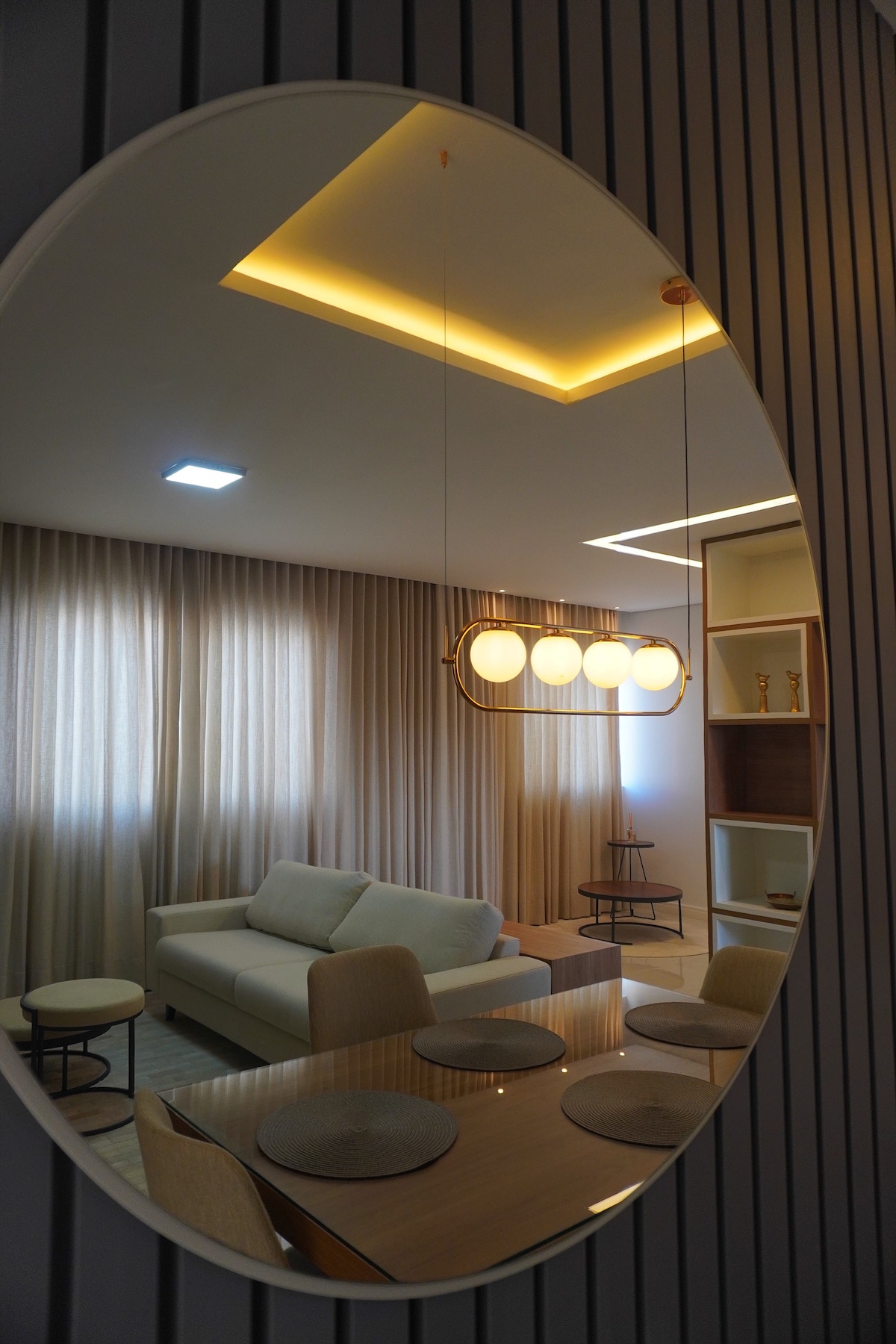 A view of a modern living space is reflected in an oval mirror. Soft lighting highlights a cozy sofa and light-colored curtains. A dining table with placemats complements a decorative shelf in the background, creating a harmonious and welcoming ambiance.